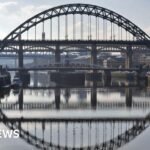 A wide view of the Tyne Bridge from the river Tyne. It is a metal bridge, painted green. Several other bridges can be seen behind it. The silhouettes of the bridges are reflected in the water below.